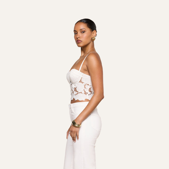 A woman stands in profile against a plain background, wearing the white Sonya Bustier crop top, white pants, gold hoop earrings, and bracelets, with her hair pulled back.