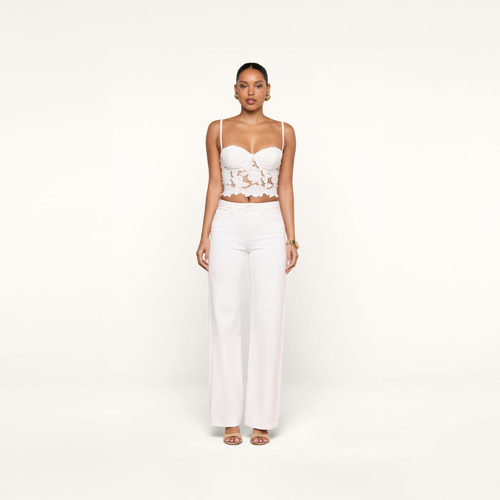 A woman stands facing forward, wearing the Sonya Bustier—a white eyelet crop top with underwire cups—paired with high-waisted white pants and sandals, against a plain white background.
