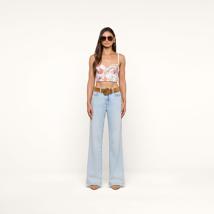 A woman with long brown hair and a neutral expression stands against a plain background, wearing sunglasses, the Sonya Bustier in a baroque toile print, light blue high-waisted flare jeans with a brown belt, and beige heels.
