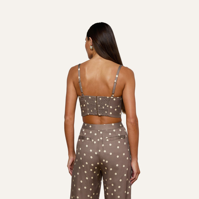 A woman with long brown hair models the Sonya Bustier—a cardamom polka dot crop top with a smocked back—paired with matching high-waisted pants, as she is seen from behind against a plain light background.