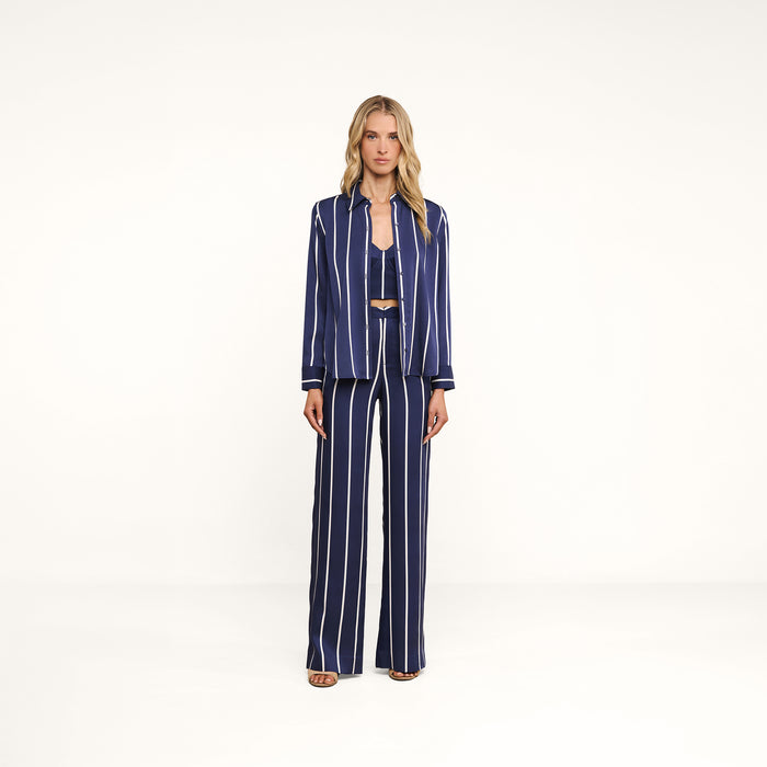 A person stands against a plain white background wearing the Livvy Trouser—a pair of high-rise, straight-leg navy blue trousers with vertical white stripes—paired with a matching button-up shirt and open-toe sandals.
