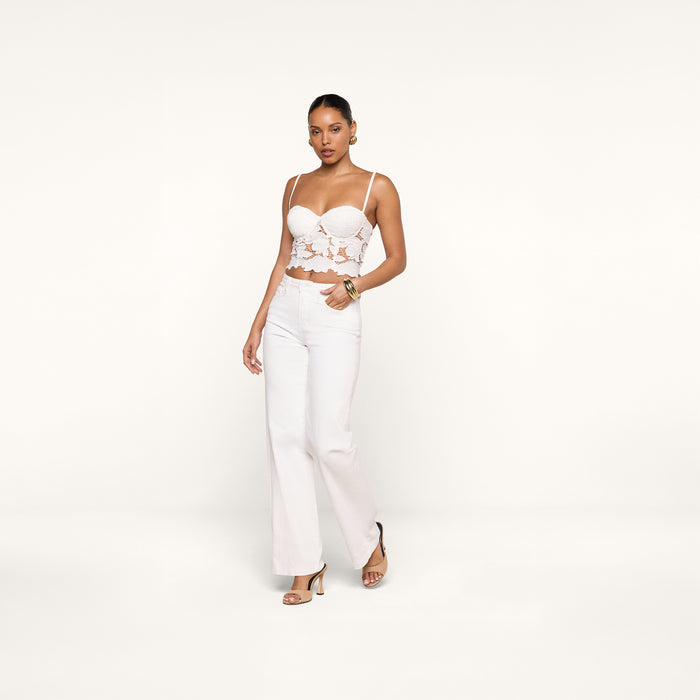 A woman poses confidently against a plain white background wearing the Sonya Bustier—a white eyelet crop top with underwire cups—paired with white high-waisted pants and heeled sandals.