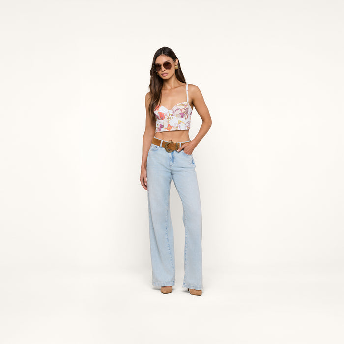 A woman wearing sunglasses, the Sonya Bustier in a baroque toile print, light blue wide-leg jeans, and beige heels stands against a plain white background.
