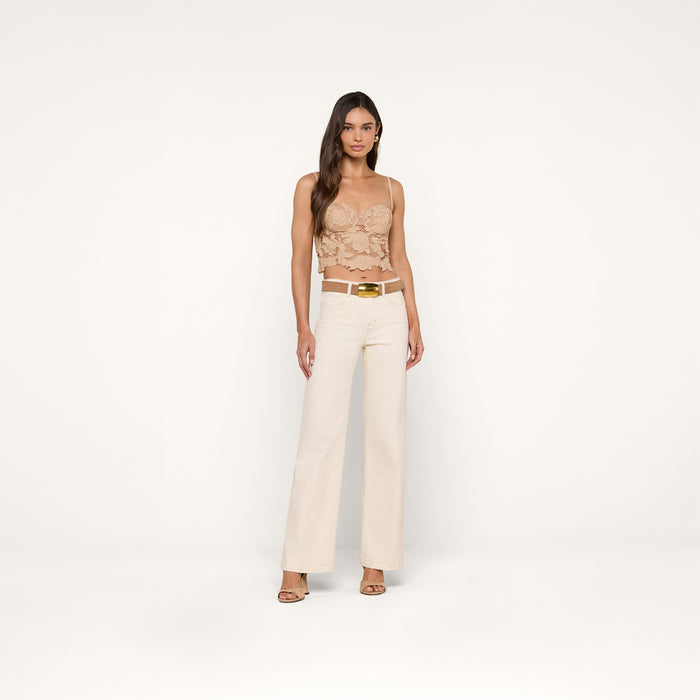 Woman standing against a plain white background, wearing the beige cotton eyelet Sonya Bustier top with adjustable straps, white high-waisted wide-leg pants, a white belt with a gold buckle, and beige heeled sandals. She has long hair and a neutral expression.