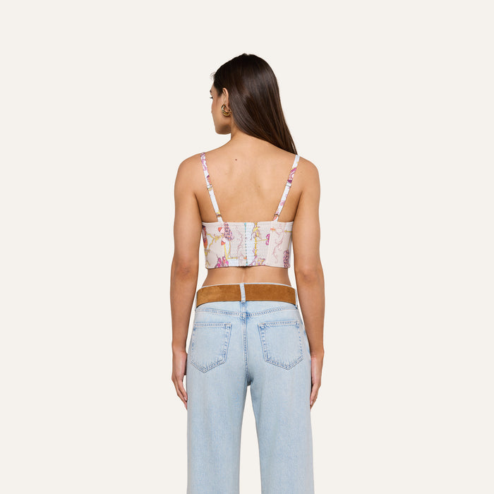 A woman with long brown hair, wearing the Sonya Bustier in a baroque toile print and light blue jeans, stands facing away from the camera against a plain light background. Her arms are relaxed by her sides.