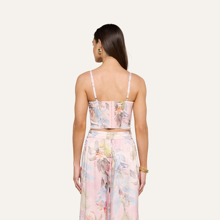 A woman stands with her back to the camera, wearing the Sonya Bustier in a pastel palm leaf print paired with matching wide-leg pants. Her straight brown hair flows down, and she accessorizes with gold hoop earrings and a gold bracelet.