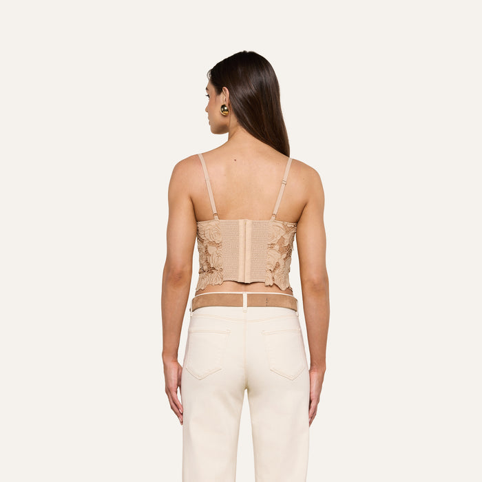 A woman with long brown hair, wearing the Sonya Bustier—a beige cotton eyelet top with adjustable straps—and high-waisted white jeans, stands facing away from the camera against a plain off-white background.