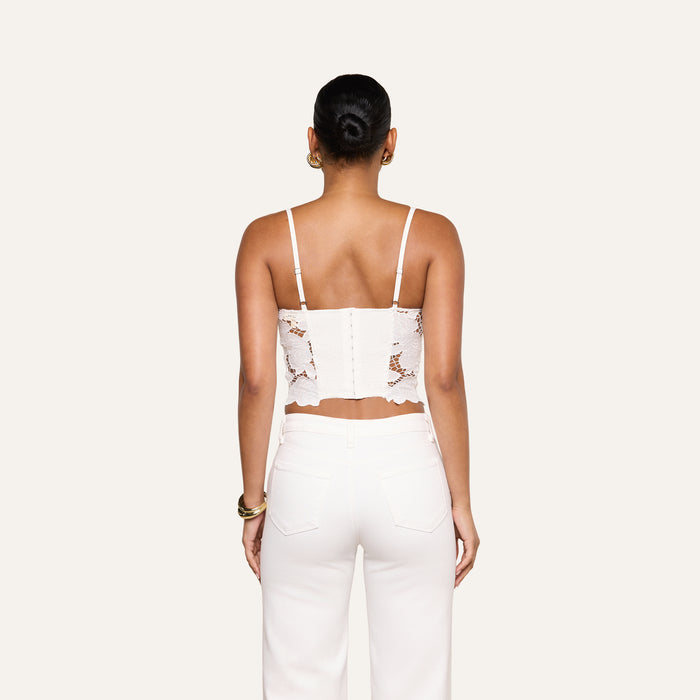 A woman stands with her back to the camera, wearing the Sonya Bustier—a white eyelet crop top with thin straps—paired with white pants. Her hair is styled in a bun, and she accessorizes with gold hoop earrings and bracelets. The background is a simple off-white.