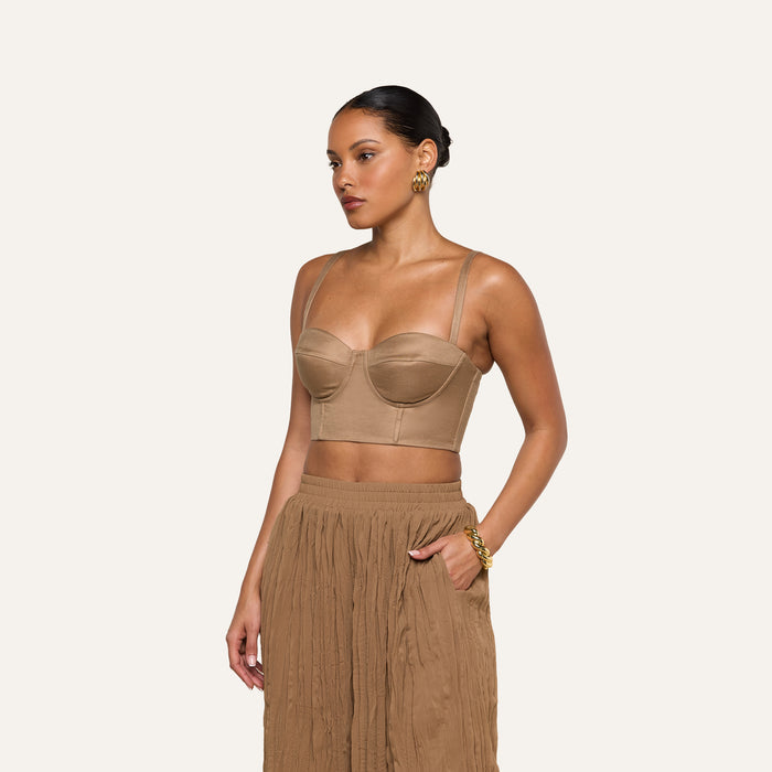 A woman stands against a plain background, looking to the side, wearing the Sonya Linen-Blend Bustier with underwire cups paired with a pleated brown skirt. Her hair is pulled back, and she accessorizes with gold earrings and a bracelet.