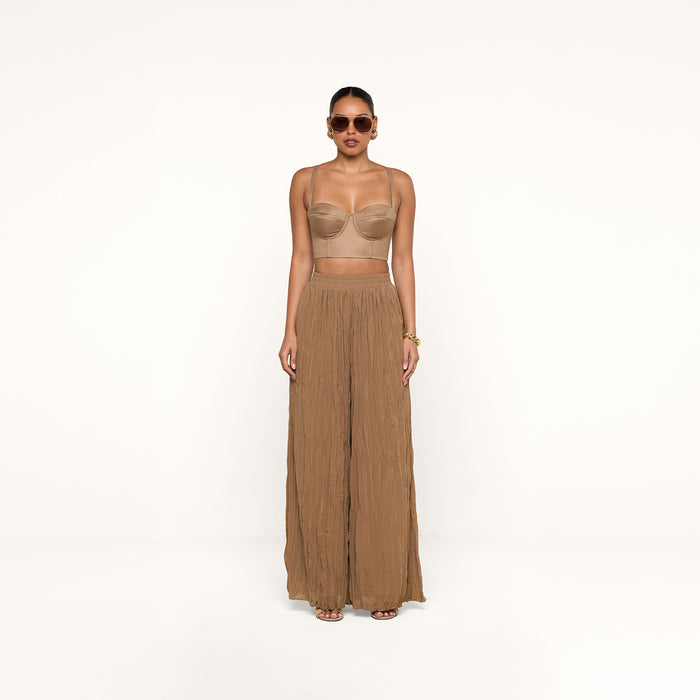A woman wearing sunglasses, the Sonya Linen-Blend Bustier with adjustable straps, and wide-legged brown pants stands against a plain white background.
