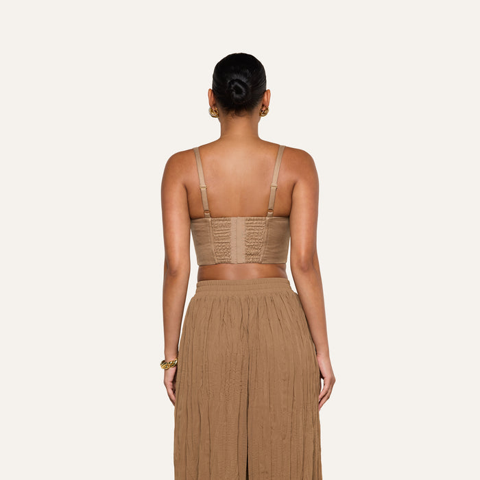 A woman stands facing away, wearing the Sonya Linen-Blend Bustier with adjustable straps and a long, pleated tan skirt. Her hair is tied in a neat bun, and the background is plain off-white.