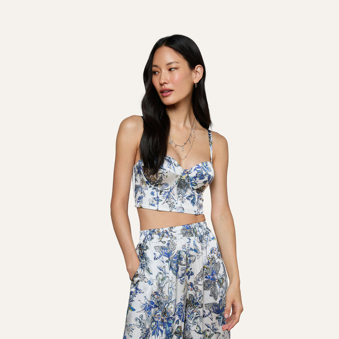 Wearing the Sonya Silk Bustier in blue and white with matching pants, a woman with long dark hair stands against a plain light background, her left hand tucked in her pocket.
