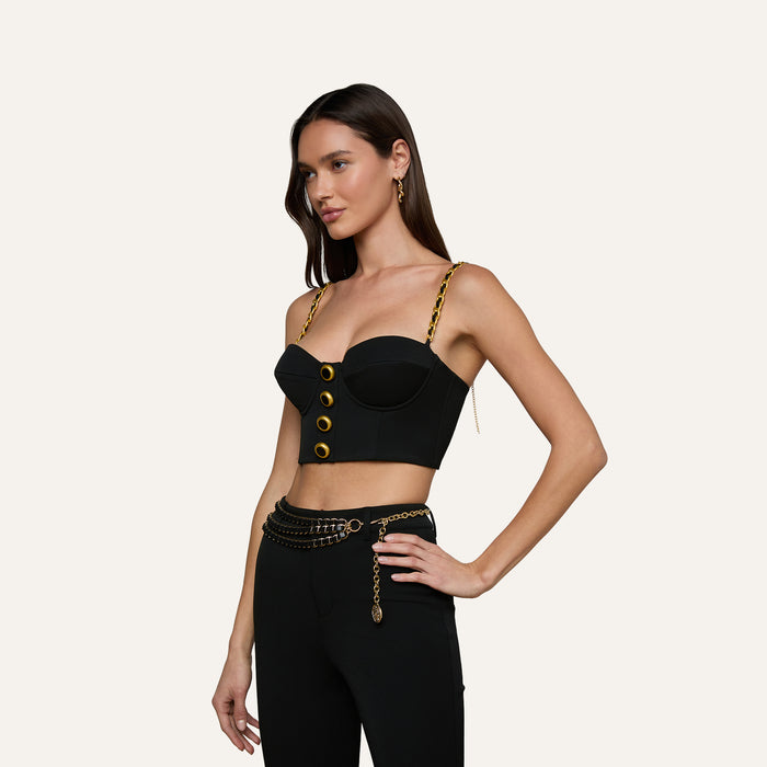 Wearing the Sophia Chain Bralette, this woman showcases a structured silhouette in a stylish black crop top with gold buttons and chain straps. She pairs it with black high-waisted pants crafted from stretch suiting fabric and completes the look with a gold chain belt, standing against a plain light background.