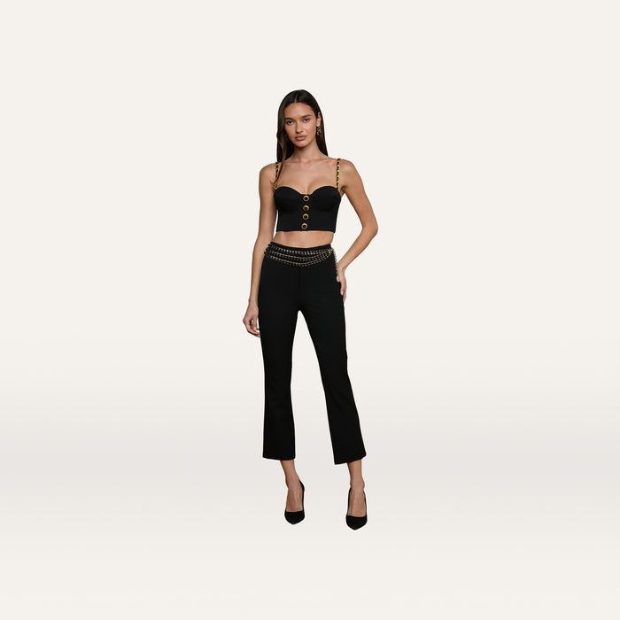 A woman wearing a black Sophia Chain Bralette with gold straps and buttons, black pants crafted from stretch suiting fabric with a gold chain belt, and black heels stands against a plain white background.