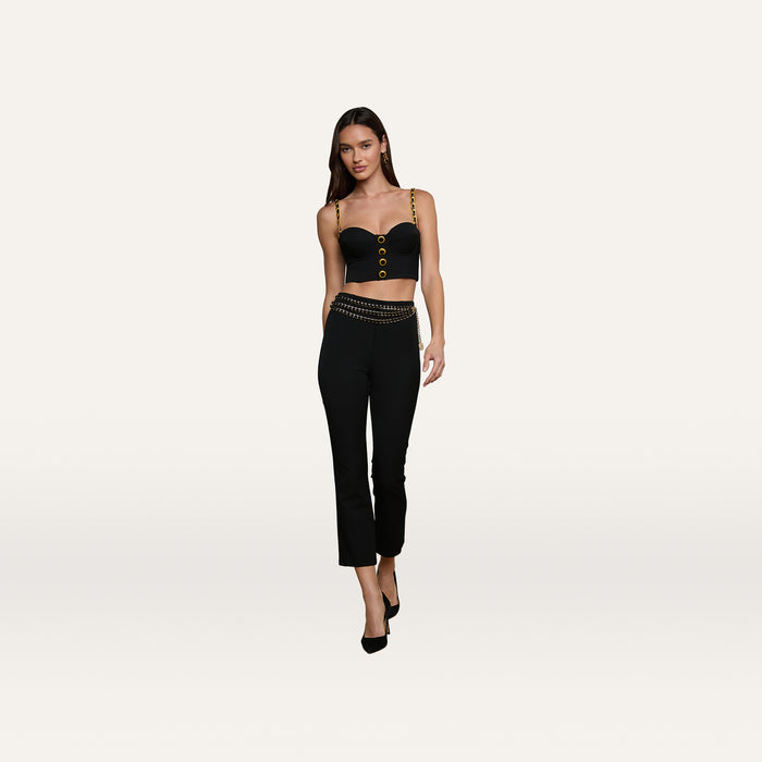 A woman wearing a black Sophia Chain Bralette with gold chain details and black pants, walking forward against a plain light background, paired with black high-heeled shoes. The outfit features a structured silhouette crafted from stretch suiting fabric.