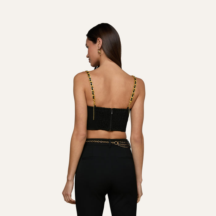 Woman with long brown hair wearing the Sophia Chain Bralette, a black crop top with gold chain straps and structured silhouette, paired with black high-waisted pants made from stretch suiting fabric, facing away from the camera against a plain light background.