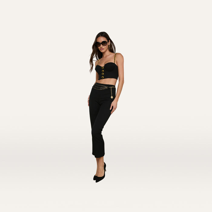 Woman wearing sunglasses, a black Sophia Chain Bralette with gold chain straps and buttons, black pants with a chain belt in stretch suiting fabric, and black heels stands against a plain white background.