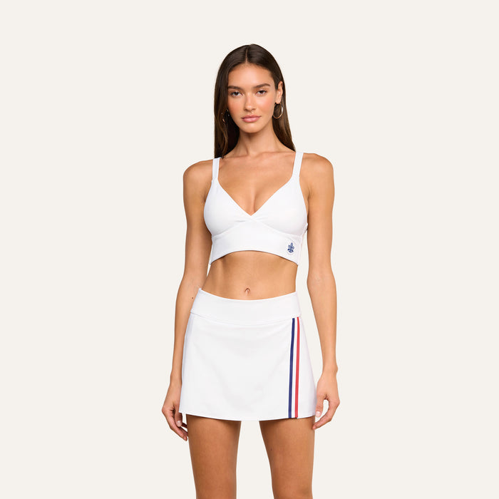 A woman wearing the Steffi Tennis Bralette and a white tennis skirt with red and blue stripes on the side, standing against a plain light background.