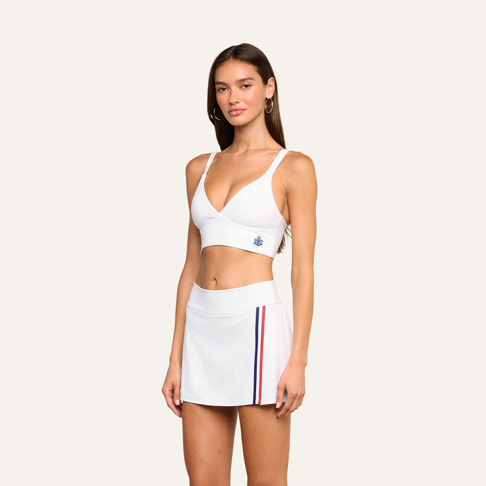 A woman wears the Steffi Tennis Bralette, a white crop top styled as a tennis bralette, paired with a white skirt featuring red and blue side stripes, standing against a plain background while looking toward the camera.