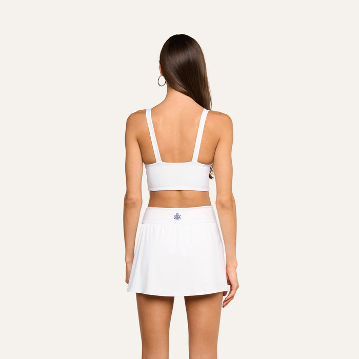 A woman with long brown hair, wearing the Steffi Tennis Bralette in white and a matching white skirt, stands facing away from the camera on a plain background.