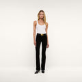 A woman stands against a plain white background wearing a white tank top, the Stevie Velvet Jean styled with a belt, and black heeled shoes. She faces forward with a neutral expression.