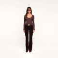 A woman wearing sunglasses, a long-sleeve dark java brown stretch velvet top, the Stevie Velvet Jean in brown with a belt, and brown shoes stands facing forward against a plain white background.