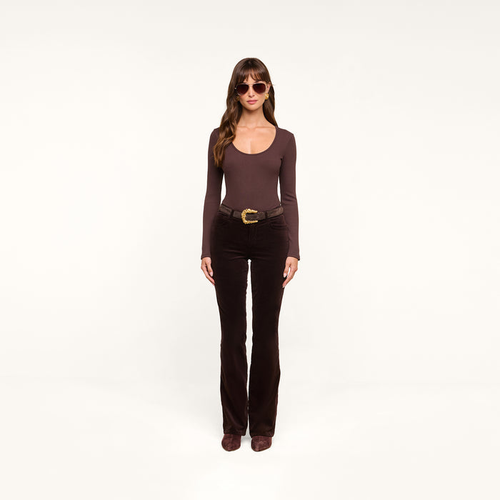 A woman wearing sunglasses, a long-sleeve dark java brown stretch velvet top, the Stevie Velvet Jean in brown with a belt, and brown shoes stands facing forward against a plain white background.