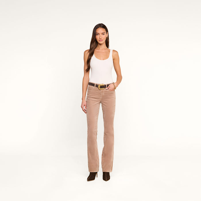 A woman stands facing forward, wearing a white tank top, the Stevie Velvet Jean with a dark belt, and black boots. The background is plain white.