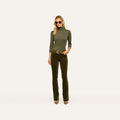 A woman wearing sunglasses, an olive green turtleneck top, and the Stevie Velvet Jean in dark green with a belt stands against a plain light background.