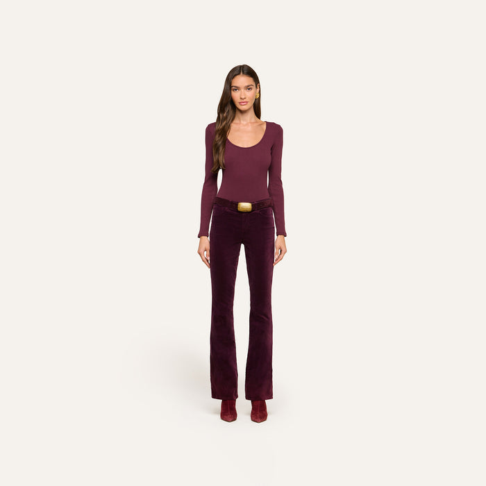 A woman stands facing forward against a plain off-white background, wearing a long-sleeve burgundy top, heeled boots, and high-rise Stevie Velvet Jeans with a belt.