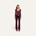 A woman stands facing forward against a plain off-white background, wearing a long-sleeve burgundy top, heeled boots, and high-rise Stevie Velvet Jeans with a belt.