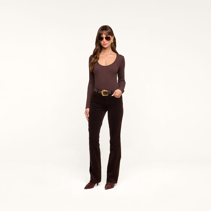 A woman wearing sunglasses, a fitted brown long-sleeve top, the Stevie Velvet Jean in slim straight leg dark java brown stretch velvet, and brown shoes stands against a plain white background.