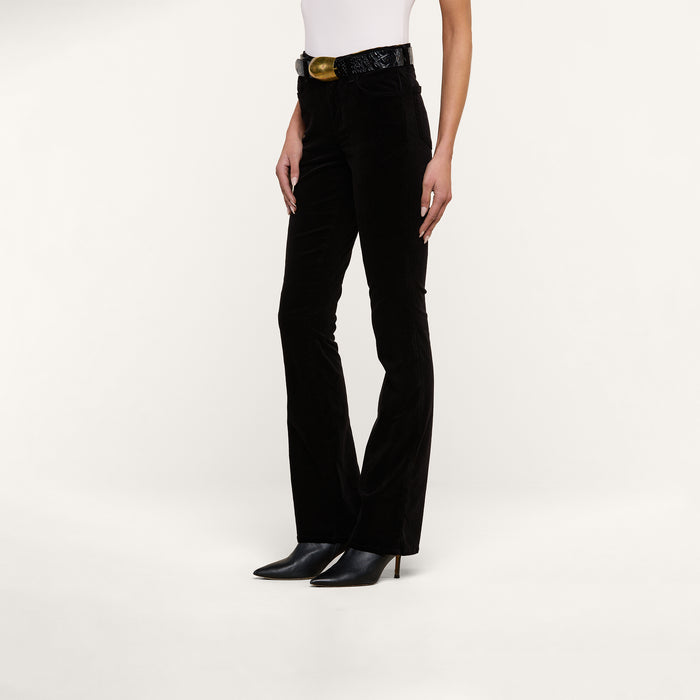 A person wearing the Stevie Velvet Jean in black, paired with a white sleeveless top, a black belt with a gold buckle, and black pointed-toe heeled boots stands against a plain white background.
