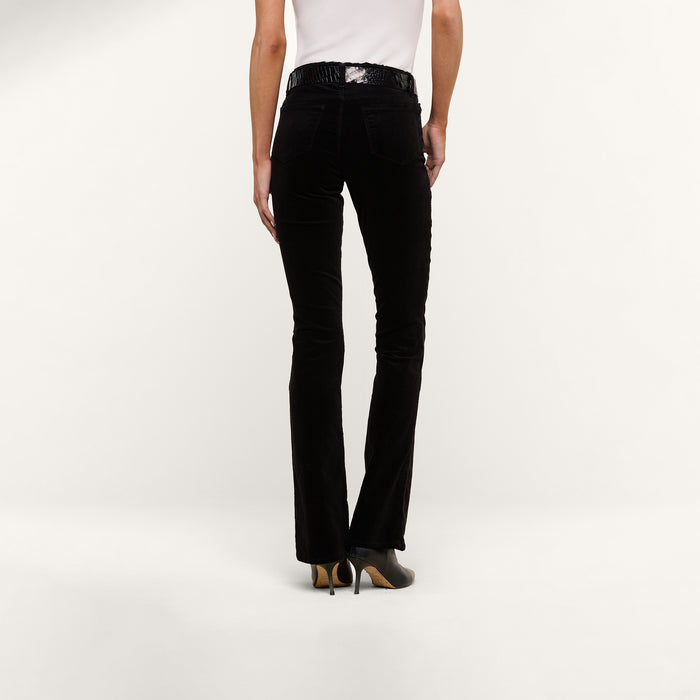 A person wearing black Stevie Velvet Jeans, a white sleeveless top, and black heeled shoes stands facing away against a plain white background.