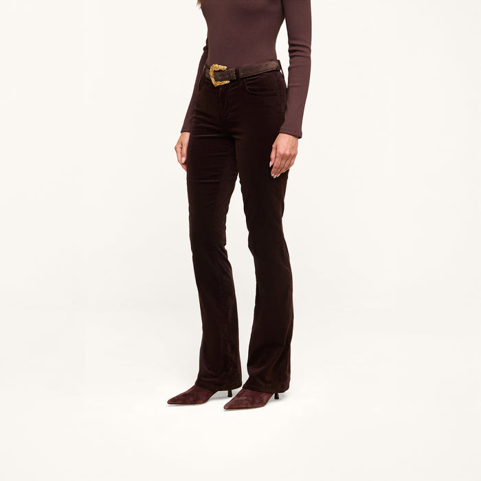 Wearing a brown long-sleeve top, the person pairs it with Stevie Velvet Jeans in a dark java brown slim straight-leg fit, accessorized with a brown belt featuring a gold buckle and pointed brown shoes. They are standing against a plain white background, with their face not visible.