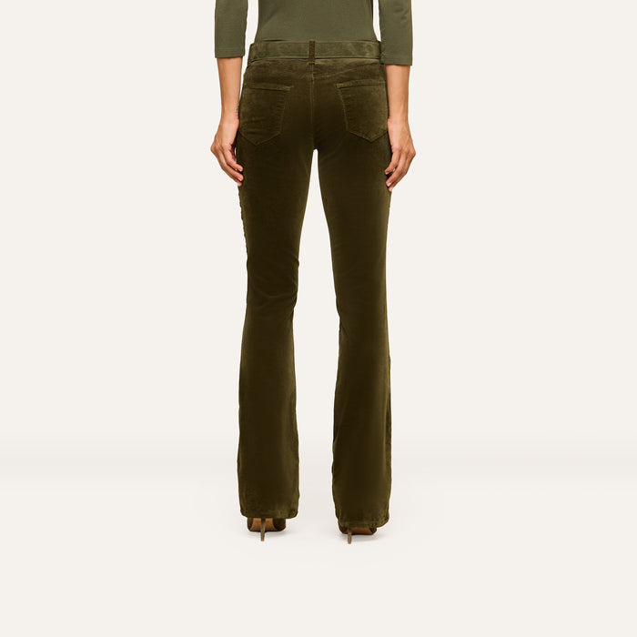 A person stands facing away, wearing the Stevie Velvet Jean—olive-green high rise pants with flared legs and back pockets—paired with heeled shoes. The background is plain and light-colored.
