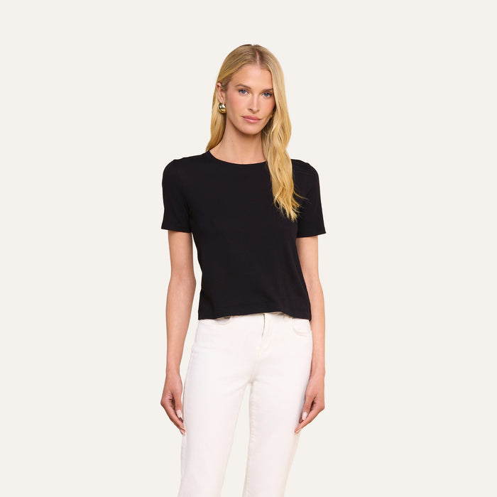 A woman with long blonde hair wears the Stormy Cropped Cotton Tee in black, featuring a semi-relaxed fit, paired with white pants as she stands against a plain light background.