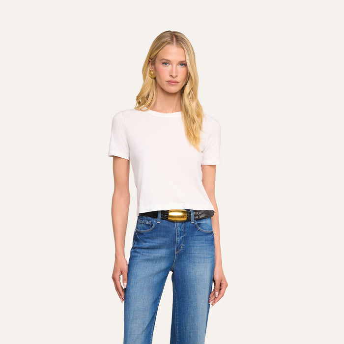 A woman with long blonde hair wears the Stormy Cropped Cotton Tee—an organic cotton short-sleeve t-shirt—in white, paired with blue jeans and a black belt with a gold buckle, standing against a light background.