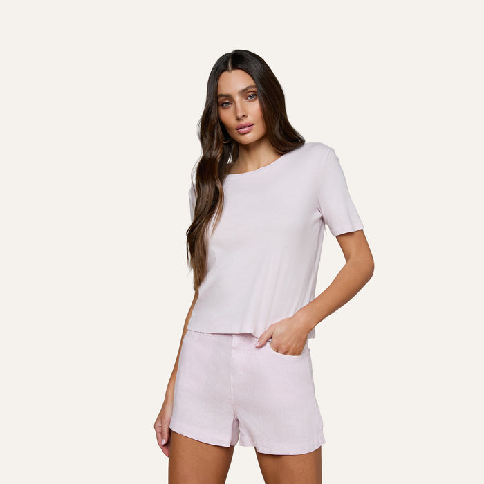 A woman with long brown hair wears the Stormy Cropped Cotton Tee in light pink—a semi-relaxed fit, cropped t-shirt—paired with matching shorts, standing with one hand in her pocket against a plain background.