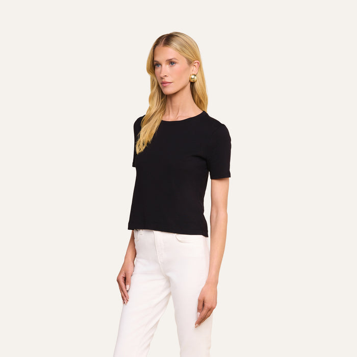 A woman with long blonde hair wears the Stormy Cropped Cotton Tee in black, featuring a semi-relaxed fit, paired with white pants. She stands against a plain light background, looking slightly to the side with a neutral expression.