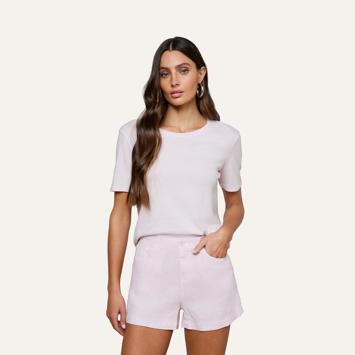 A woman with long wavy brown hair stands against a plain background, wearing the Stormy Cropped Cotton Tee in light pink and matching shorts, with one hand tucked into her pocket.