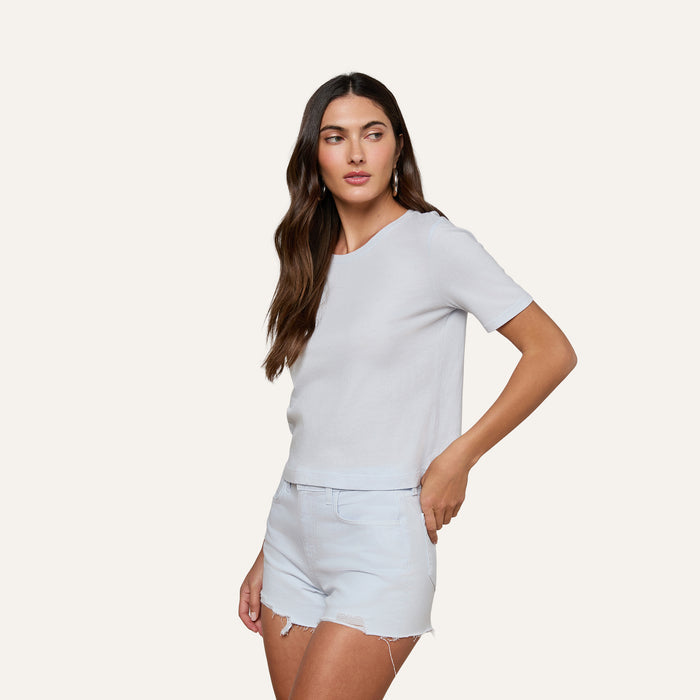 A woman with long brown hair wears the Stormy Cropped Cotton Tee, a plain white organic cotton shirt in a semi-relaxed fit, paired with light denim shorts. She stands against a plain light background, gazing slightly to the side with one hand near her shorts pocket.