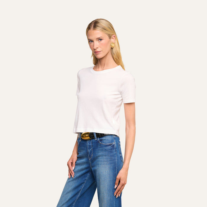 A person with long blonde hair wears the Stormy Cropped Cotton Tee in white, styled in a semi-relaxed fit with blue jeans, a black belt featuring a gold buckle, and large round earrings, posing against a plain light background.