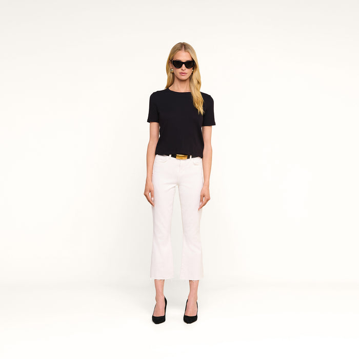 A woman wearing the Stormy Cropped Cotton Tee in black, paired with sunglasses, white pants, and black heels stands against a plain white background.