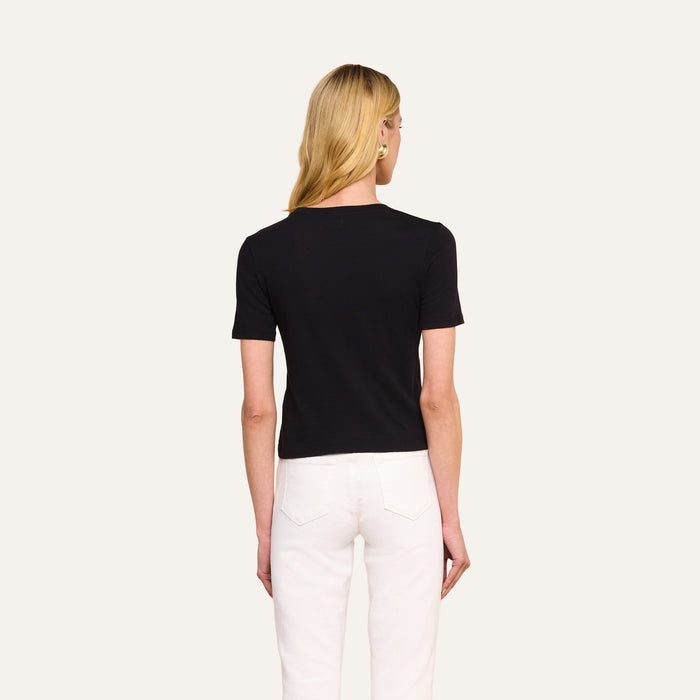 A person with long blonde hair, wearing the Stormy Cropped Cotton Tee in black—a semi-relaxed fit organic cotton top—and white pants, stands facing away from the camera against a plain, light background.