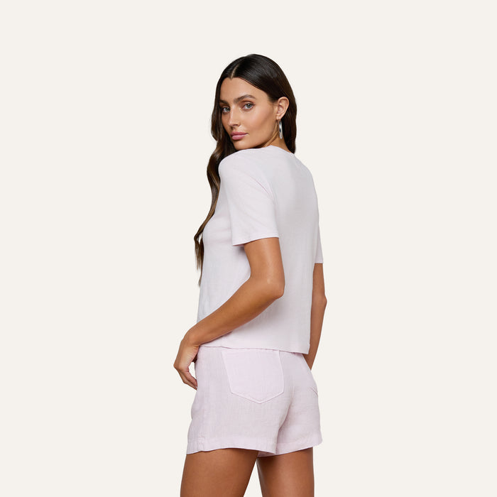 A woman with long brown hair wears the Stormy Cropped Cotton Tee in light pink paired with matching shorts, standing against a plain light background and looking over her shoulder with one hand on her hip.