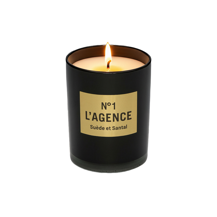 A lit Suède et Santal black sandalwood candle, crafted from soy-blend wax and housed in a glass jar with a beige label reading “No 1 LAGENCE Suède et Santal,” sits on a white background.