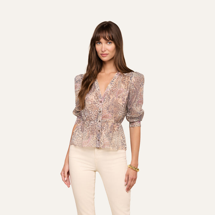 A woman with long brown hair wears the Suri Top with a paisley animal print and puff sleeves, paired with cream pants. She stands against a plain light background, looking at the camera with a slight smile.