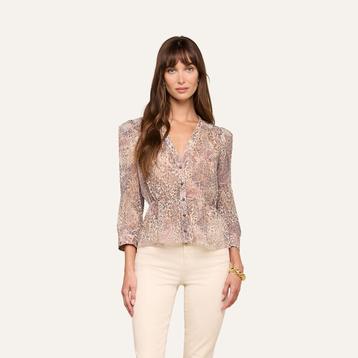 A woman with long brown hair wears the Suri Top—a sheer poly chiffon blouse featuring puff sleeves and a paisley animal print—paired with light-colored pants, standing against a plain light background.
