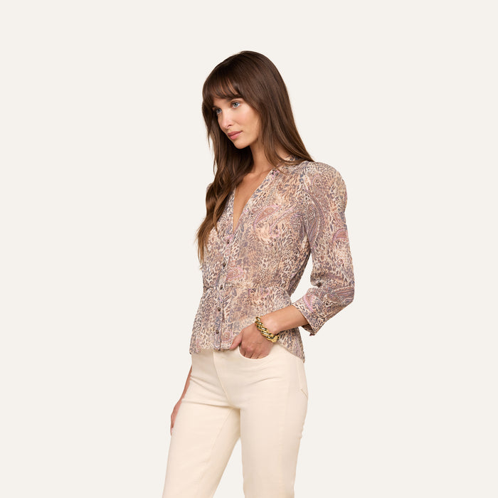 A woman with long brown hair and bangs stands sideways against a plain background, wearing the Suri Top—a long-sleeved poly chiffon blouse with a paisley animal print and V-neck—paired with light-colored pants. She has one hand in her pocket.